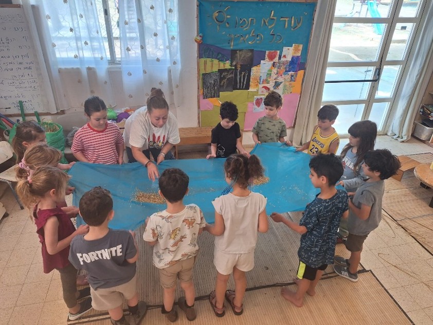 ילדי הגן עומדים במעגל ומניפים יחד מצנח בד כחול עם זרעים, בפעילות קבוצתית בתוך הגן