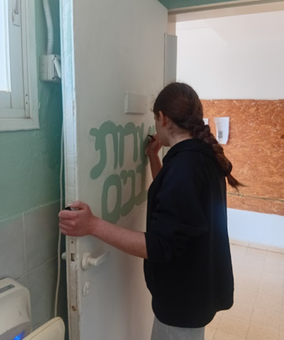 תלמידה מתנדבת צובעת כתובת בעברית על דלת, במסגרת פעילות התנדבות בבית הספר