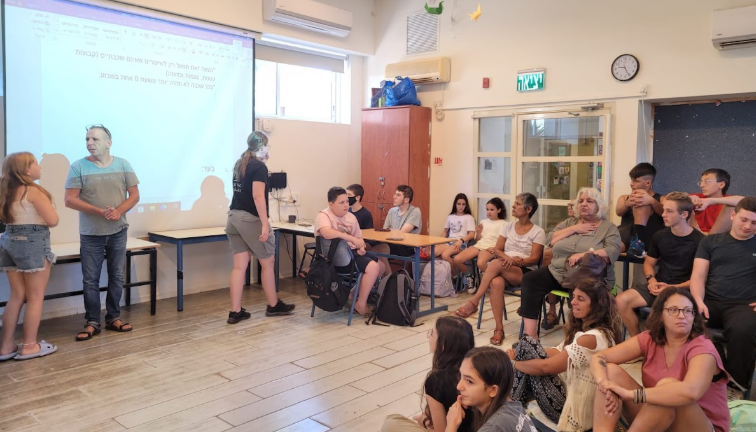 תמונה ממפגש פרלמנט בית הספר: תלמידים והורים יושבים ומקשיבים למציג לצד מצגת